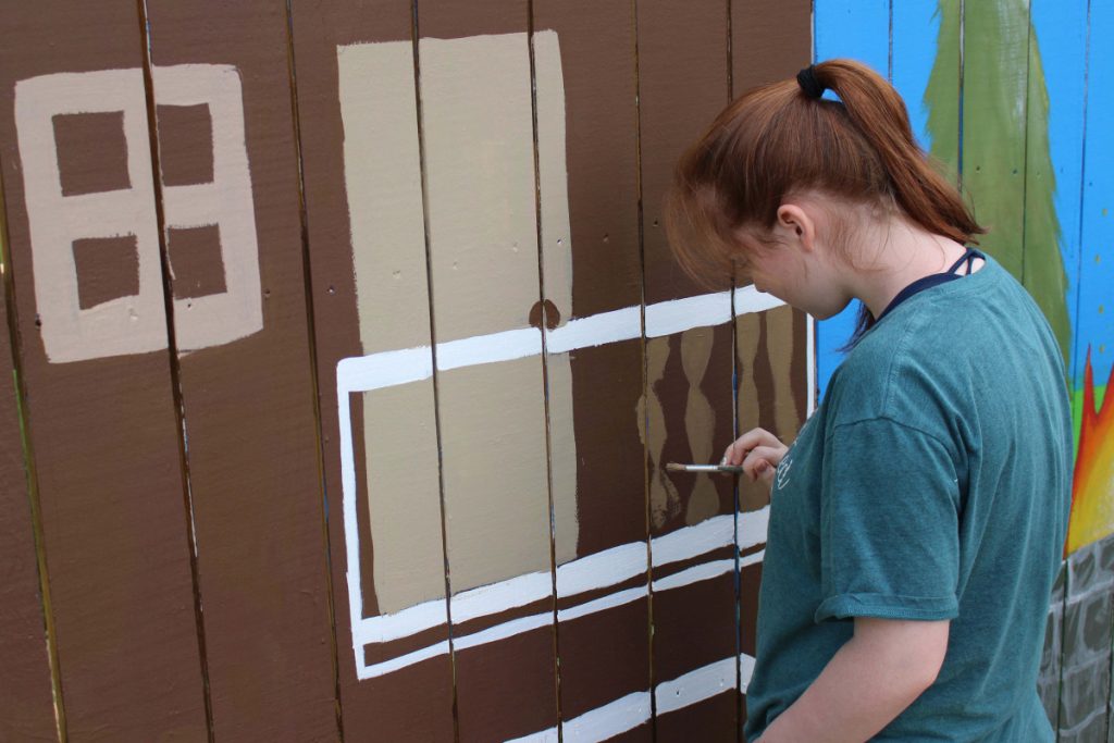 Student paints a mural on a  wooden fence. 
