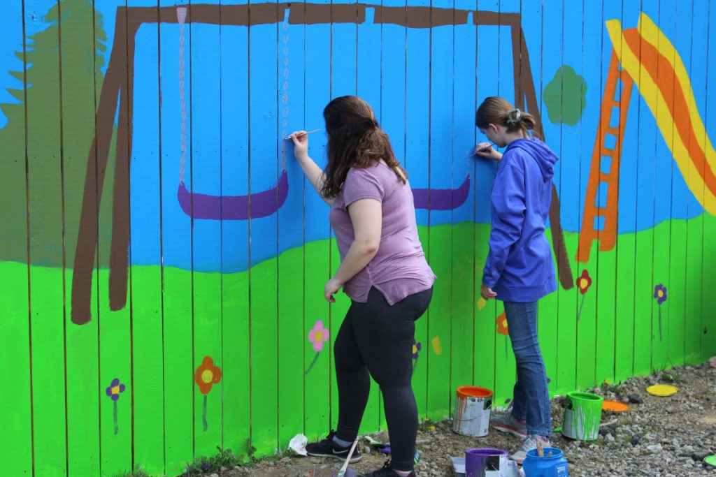 Students paints a mural on a  wooden fence. 